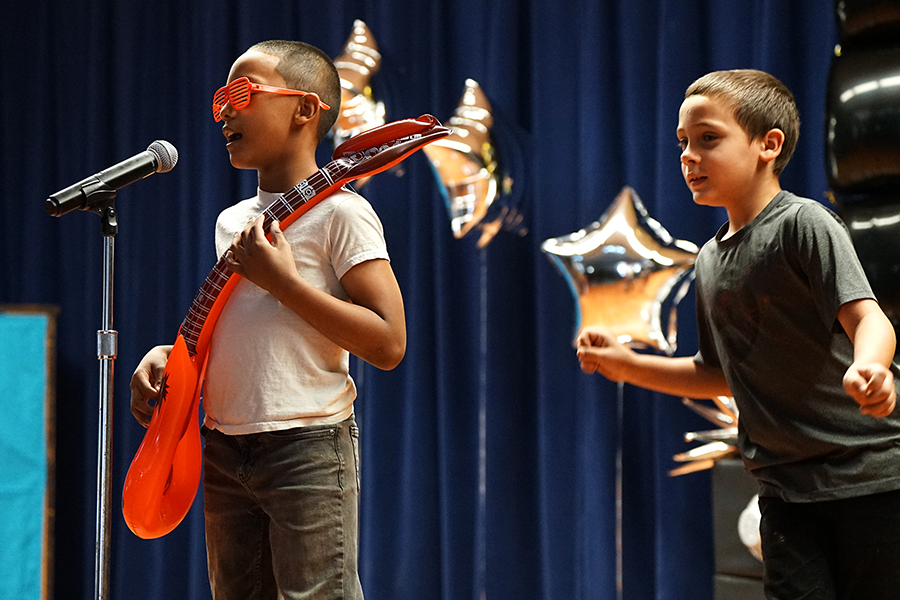 A boy in orange sunglasses plays an orange inflatable guitar, while another boy dances behind him