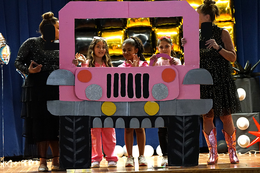 A group of girls perform a dance behind a pink bus cutout