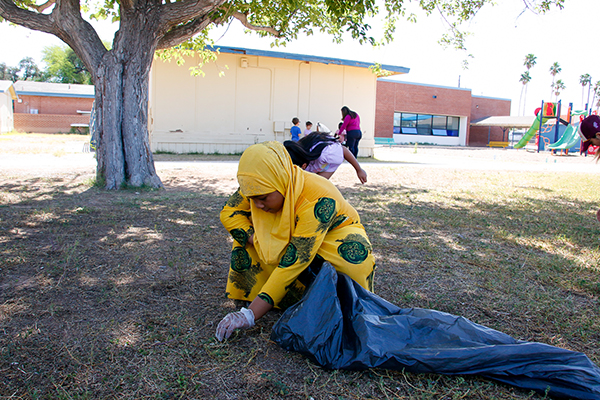 A girl in a yellow dress and headscarf leans over to pick up trash