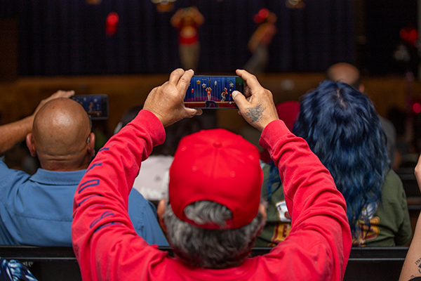 A man in a red sweatshirt holds up his phone to take a video of the kids performing on stage