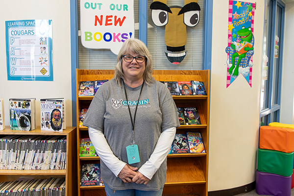 A woman in a gray shirt and glasses smiles in front of a shelf of books